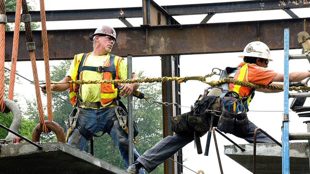 A construction worker stretching to a beam while attached by a rope.